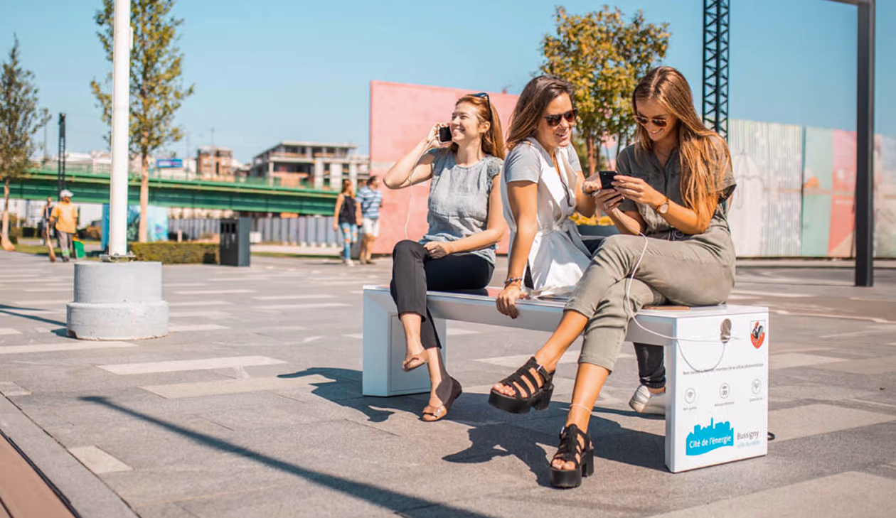 Three women sitting on SSB smart bench outdoors using phones